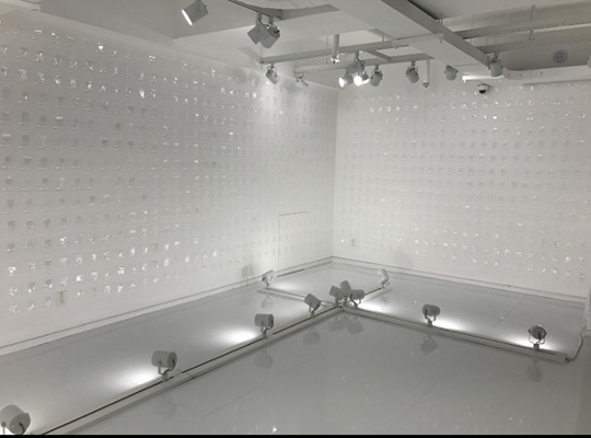 Corner view of a white gallery installation with transparent wall-mounted elements and floor lights