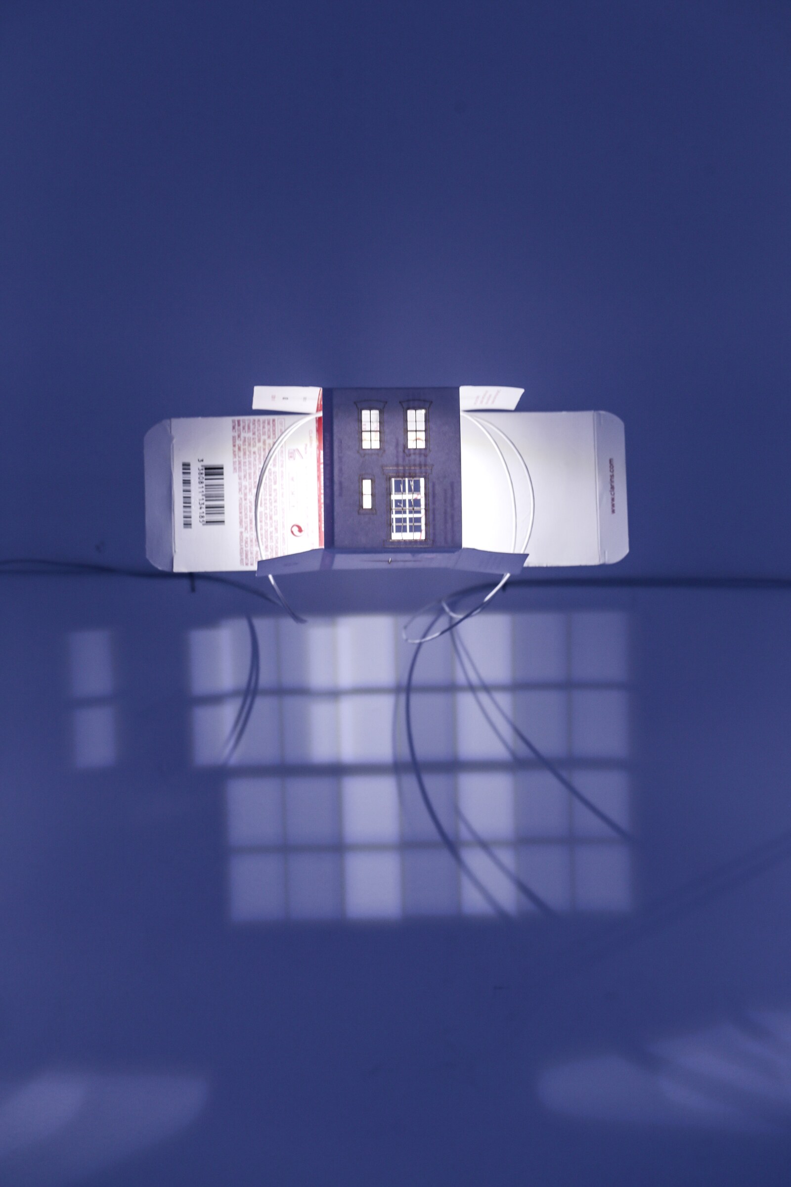 Illuminated house-like architectural form casting a window-grid shadow in a dark gallery